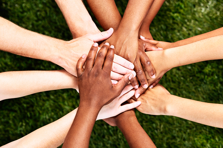 A pile of  multiracial hands are stacked in support or unity, against a background of grass. All for one and one for all!